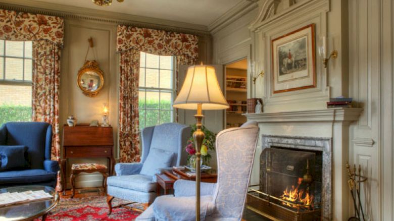 A cozy living room with a lit fireplace, floral curtains, two armchairs, a table lamp, and a mirror over a small desk, creating a warm ambiance.