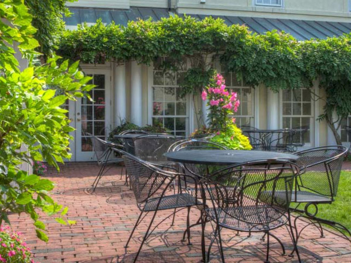 A patio with several black metal tables and chairs surrounded by lush greenery next to The Francis Malbone House.