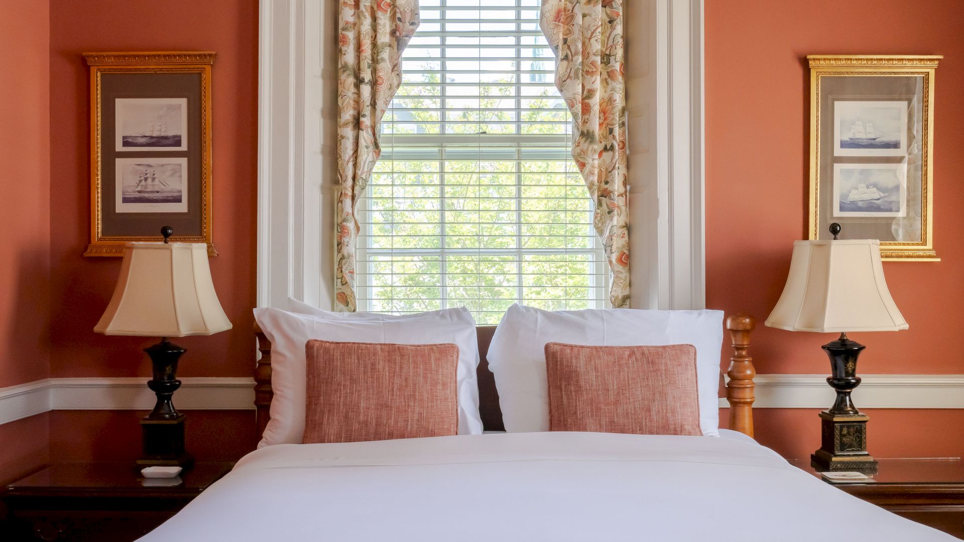 A cozy Newport, RI, guest room with a queen-size bed with white linens and red decorative throw pillows with sidetables and lamps.