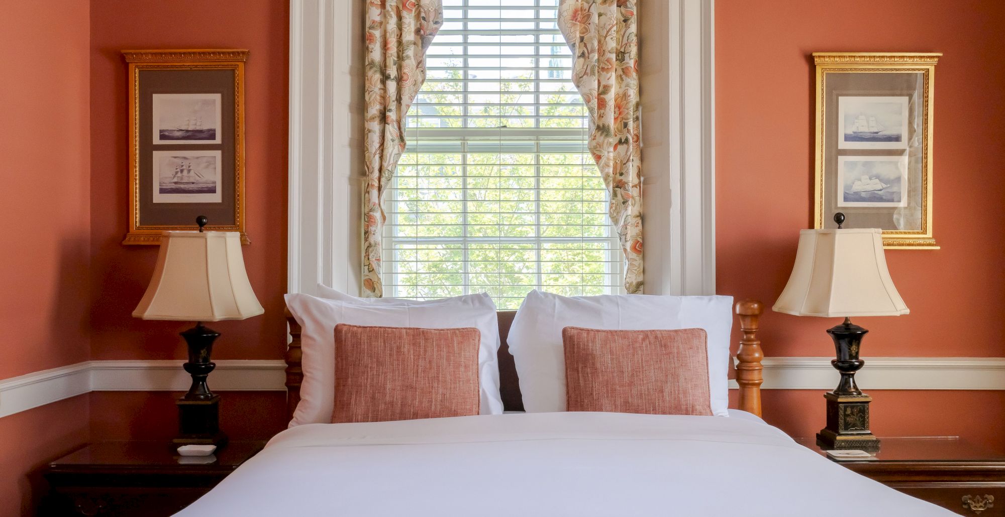 A cozy Newport, RI, guest room with a queen-size bed with white linens and red decorative throw pillows with sidetables and lamps.