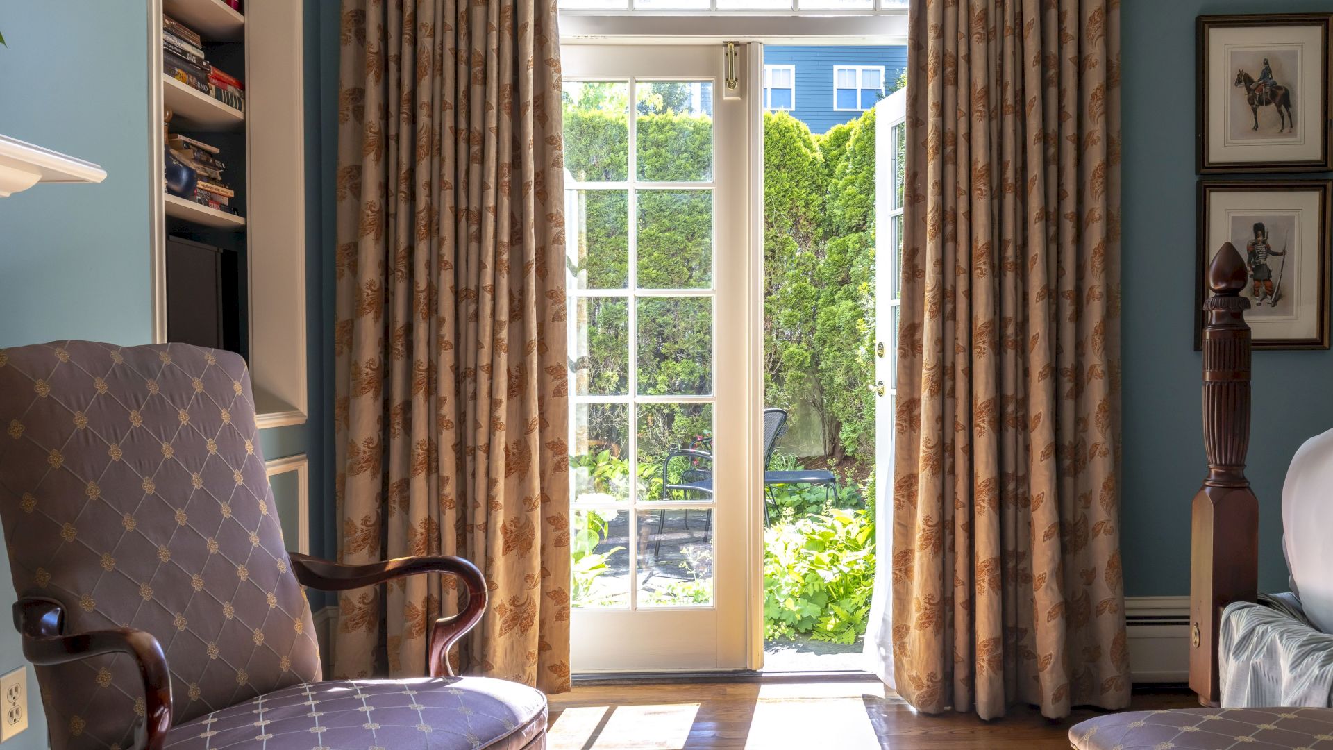 A cozy Newport, RI, guest room with an armchair and French doors overlooking a shared patio with lush greenery.