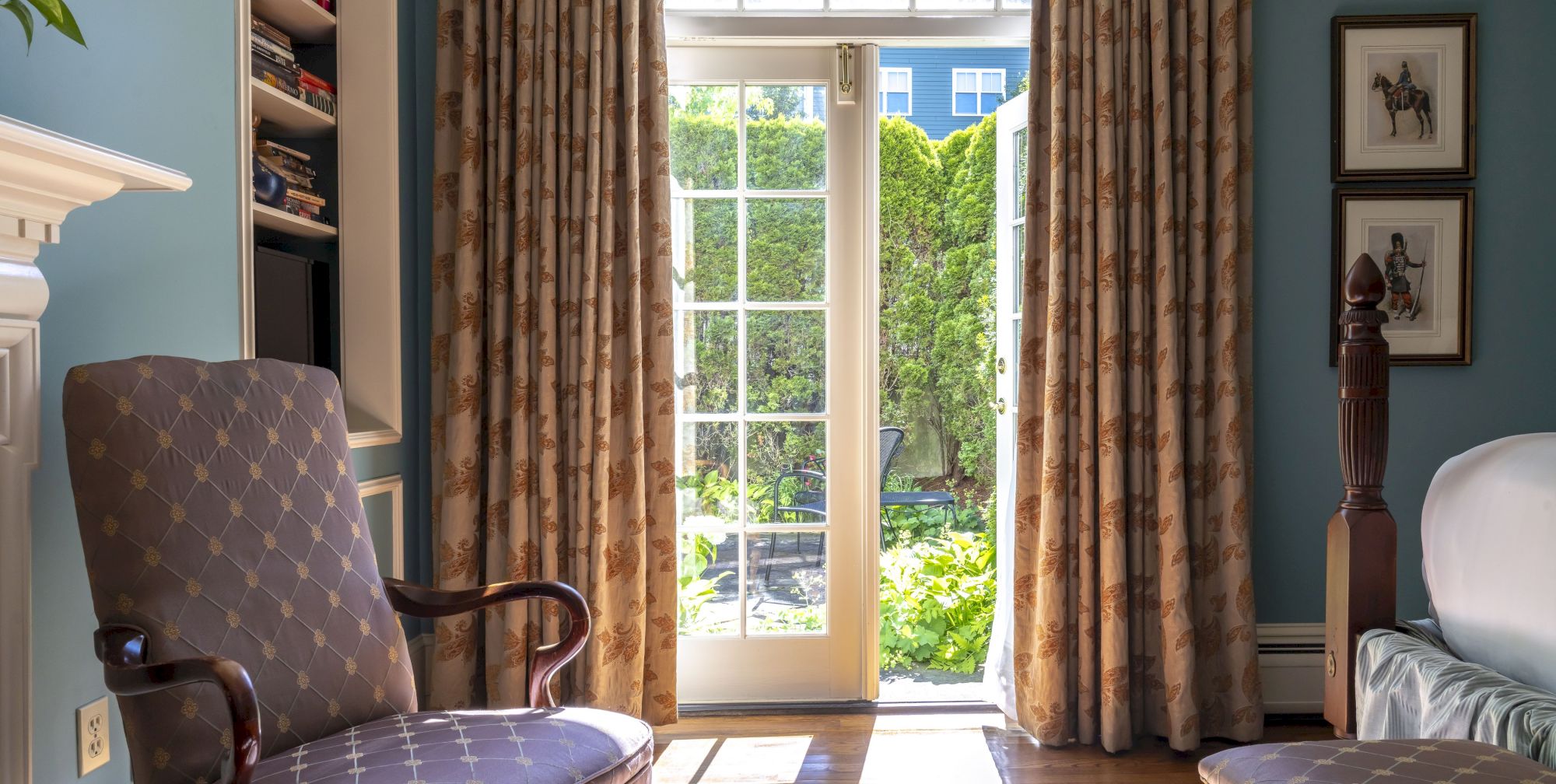 A cozy Newport, RI, guest room with an armchair and French doors overlooking a shared patio with lush greenery.