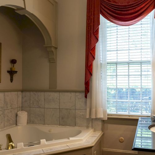A bathroom featuring a jacuzzi tub, a vanity sink with toiletries, a large window with red curtains, and a decorative wall piece.