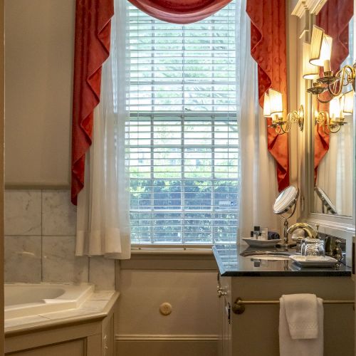 A bathroom with a sink, mirror, and bathtub. It features a red curtain, wooden flooring, and a window with blinds.
