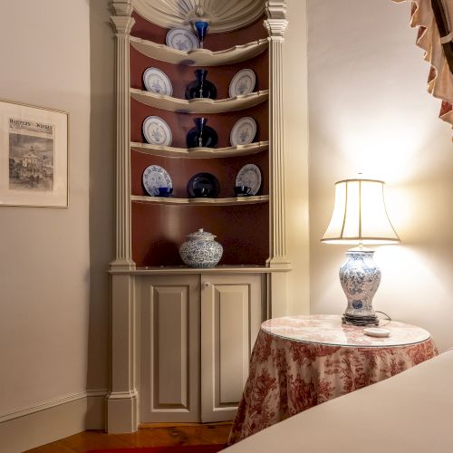 A corner cabinet displays decorative blue and white plates and vases. A nearby lamp is on a table with a patterned cloth.