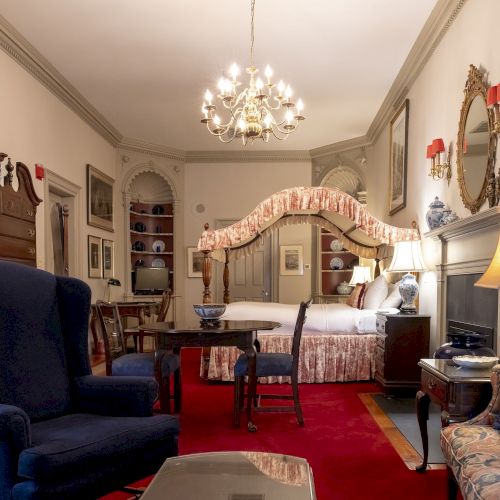 A luxurious room with vintage decor, featuring a canopy bed, blue armchair, ornate chandelier, and classic artwork on the walls.