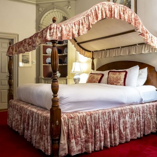 A luxurious, elegant bedroom with a canopy bed, adorned in floral fabric, and classic furnishings against a red carpet backdrop.