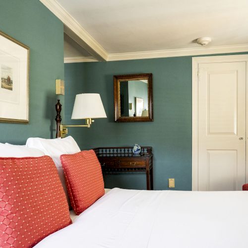 A guest room with two red pillows, white bedding, a framed picture, a mirror, and a red armchair against teal walls and a door.