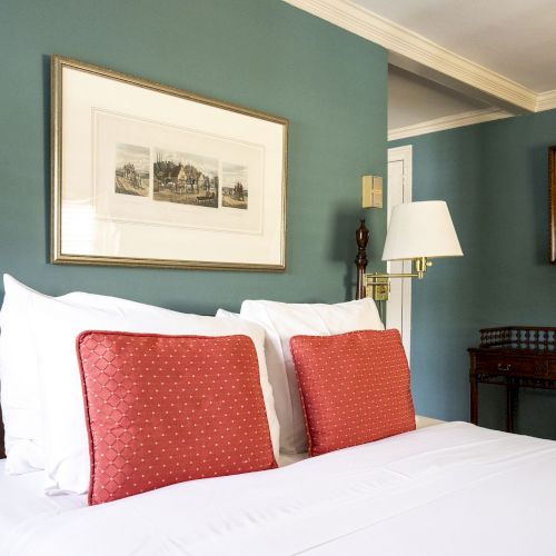 A neatly made bed with red cushions, teal walls, art, a wall lamp, mirror, and wooden side table with a blue vase.