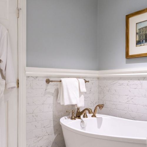 A bathroom with a white robe, towel on a rack, bathtub with bronze fixtures, tiled wall, and framed painting on a gray wall.