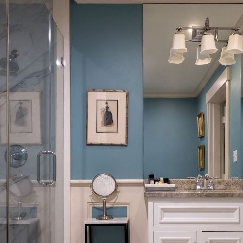 A bathroom with a marble shower, blue walls, a vanity mirror, framed art, and a countertop with toiletries and glassware.