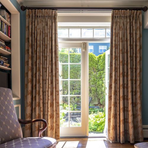 A cozy room with blue walls, armchair, books, curtains, and an open door to a garden, allowing sunlight in. Air conditioner above.