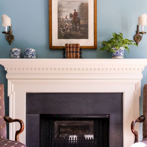 A fireplace with a framed painting, antique books, and potted plants on the mantel, flanked by chairs and wall sconces.