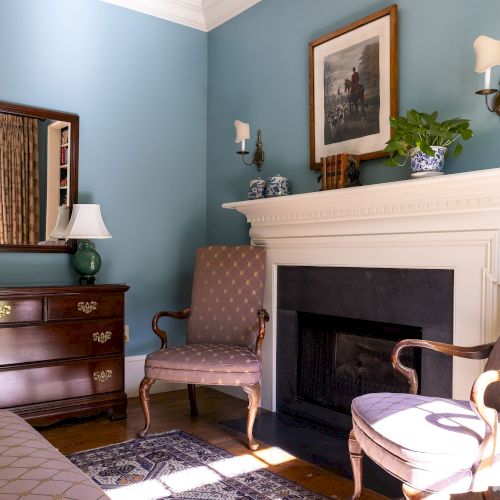 A cozy room with blue walls features a fireplace, armchairs, a dresser with a lamp, and a mirror reflecting curtains and bookshelves.