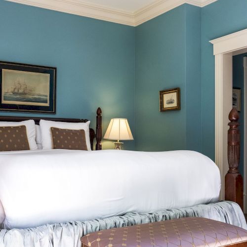 A bedroom with a four-poster bed, blue walls, framed ship artwork, a bedside lamp, and a padded bench at the foot of the bed.