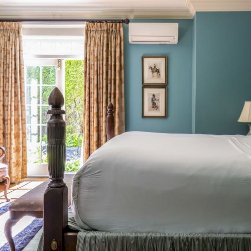 A cozy bedroom with a large bed, elegant armchair, bookshelves, art, and curtains framing a bright window leading outdoors.