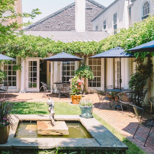 The image shows a charming courtyard with a fountain, potted plants, tables, and blue umbrellas, surrounded by lush greenery and a building in the background.