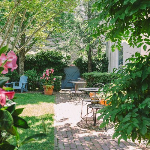 A garden with blooming flowers, brick pathway, outdoor seating, and lush greenery surrounding a cozy sitting area.