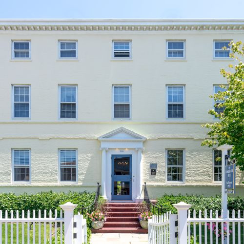 The image shows a three-story yellow building with a white picket fence, multiple windows, a central door with red steps, and surrounding greenery.