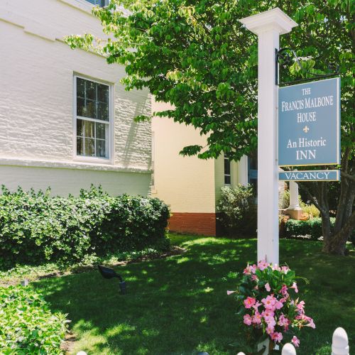 The image shows a garden with green grass, bushes, a tree, flowers, and a sign for 