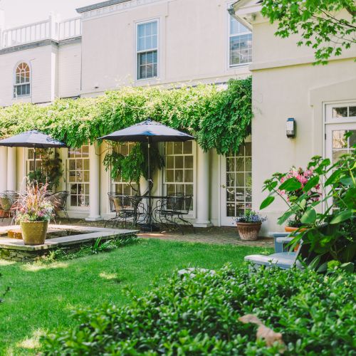 A charming garden courtyard with lush greenery, patio furniture under umbrellas, colorful potted plants, and a picturesque building facade in the background.