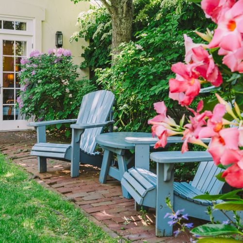 A cozy garden scene with blue Adirondack chairs, a table, vibrant pink flowers, lush greenery, and a pot of flowers near a door.