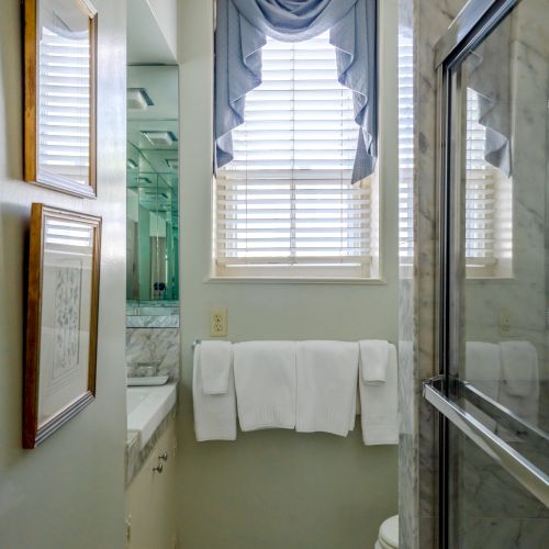 A small bathroom with a window, patterned curtains, a towel rail with two white towels, framed pictures, a shower with sliding glass doors, and a white hex tile floor.