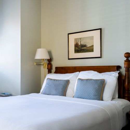 A neatly made bed with white linens and blue pillows, flanked by two bedside lamps, and a framed picture above the headboard in a well-lit room.