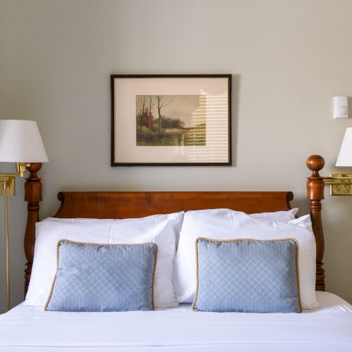 The image shows a neatly made bed with white linens, two blue pillows, wall lamps, and a framed picture above the headboard.