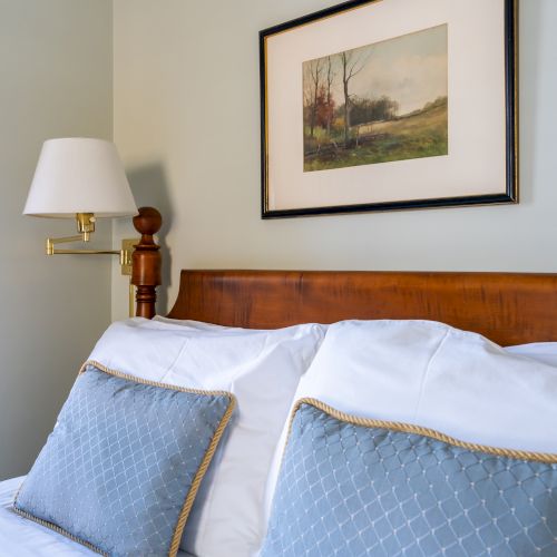 A bed with blue pillows, white sheets, a wooden headboard, a wall-mounted lamp, and a framed landscape painting above it.