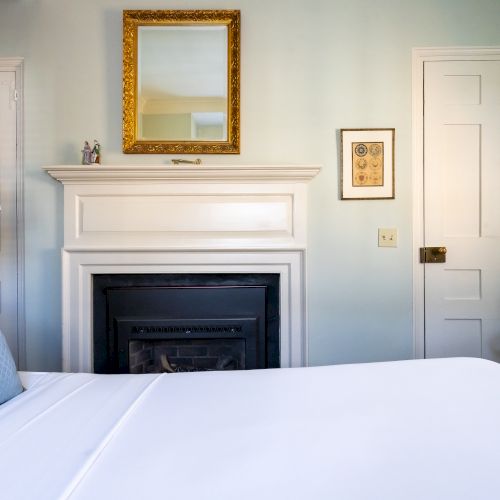 A cozy bedroom features a neatly made bed, a fireplace with a gold-framed mirror above, a chair, and two closed doors in soft, light tones.