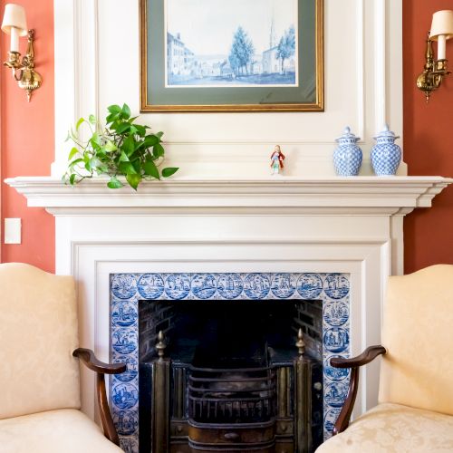 A cozy fireplace with a decorated mantel, featuring a framed painting, two blue vases, a plant, and two cream-colored chairs.