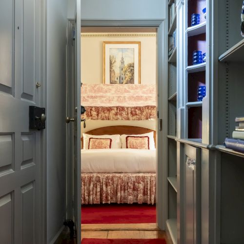 A narrow, carpeted hallway with built-in shelves leads to a bedroom with a neatly made bed and framed artwork on the wall.