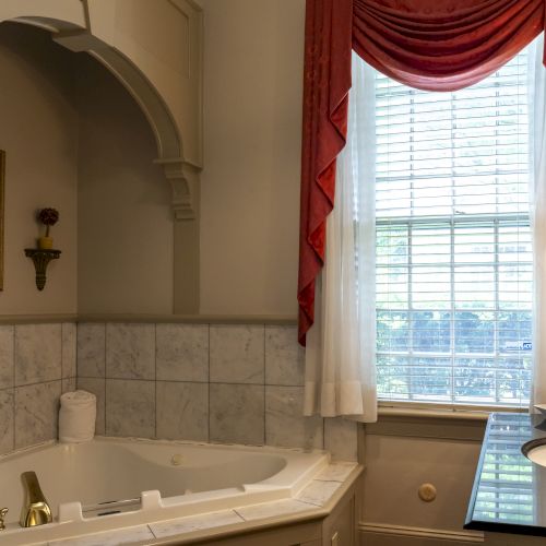 A bathroom with a corner bathtub, tiled walls, a window with red curtains, and a vanity sink area with a mirror and light fixtures.