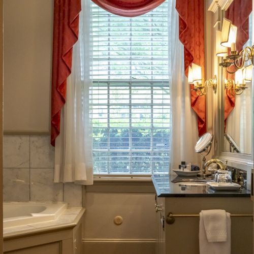 This image shows a bathroom with a bathtub, a vanity with a sink, a mirror, and a window with red and white curtains.