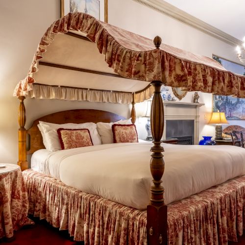 A luxurious bedroom with a canopy bed, elegant lamps, a fireplace, ornate furnishings, and a chandelier, featuring warm, rich decor and a red carpet.