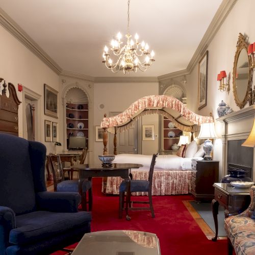 The image shows a well-furnished, elegant bedroom with a chandelier, a canopy bed, armchairs, a fireplace, and classic decor.