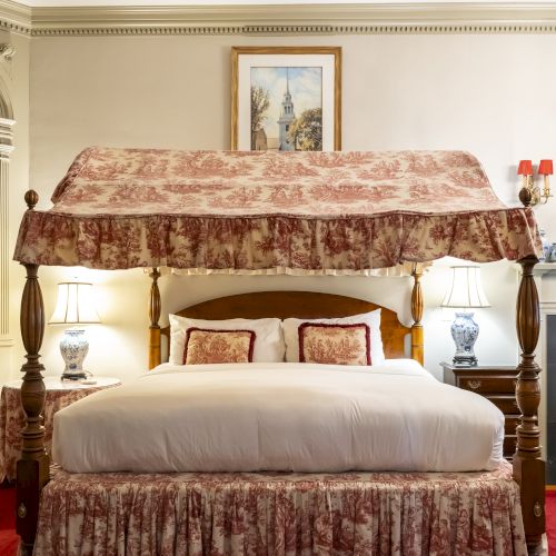 A four-poster bed with floral canopy and bed skirt, flanked by two bedside tables with lamps, in an elegant room with framed wall art.