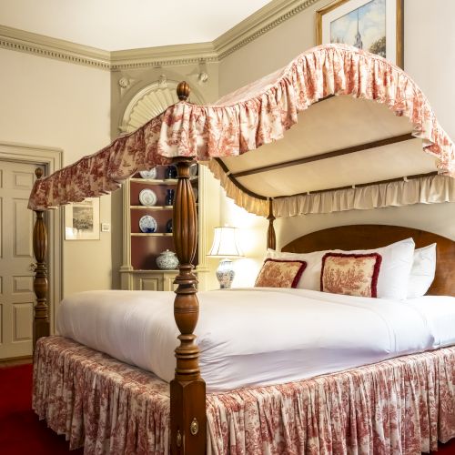 The image shows a bedroom with a canopy bed adorned with floral fabric, matching bed skirt, pillows, red carpet, and elegant decor on the walls and shelves.