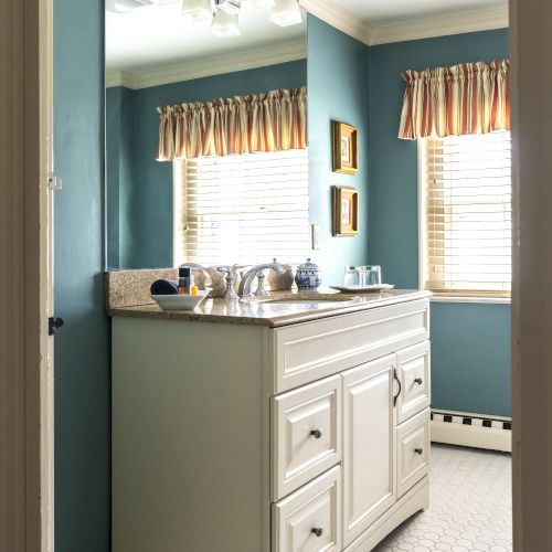 A bathroom with a white vanity, blue walls, and windows with beige curtains. The countertop has toiletries, and the floor is tiled.