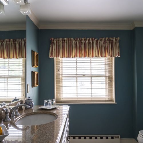 A bathroom featuring a granite countertop with a sink, mirror, and toiletries. Two windows with blinds and valances are visible, along with a shower curtain.