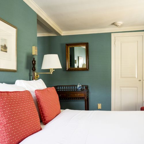 A cozy bedroom with teal walls, a white bed, red pillows, a red armchair, a framed artwork, a mirror, and a wooden side table with a lamp.