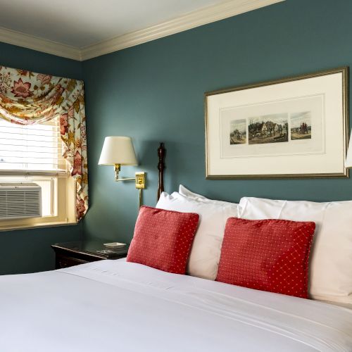 A neatly made bed with white linens and red pillows in a cozy room, featuring wall lamps, a framed picture, floral curtains, and a window A/C unit.