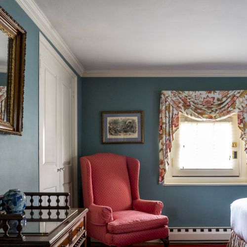 A cozy room with blue walls, a red armchair, a bed, a mirror, a desk with a blue vase, and a window with floral curtains.