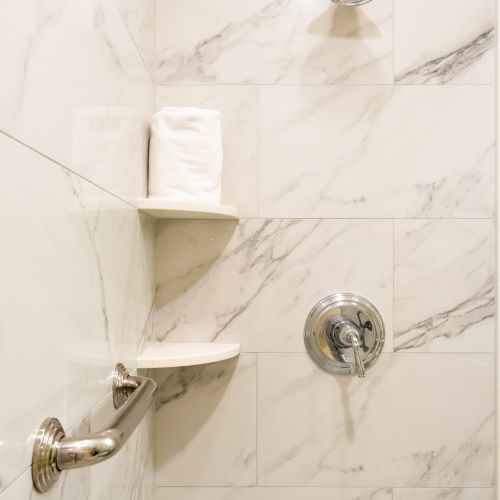 A marble-tiled shower with a showerhead, a grab bar, a control knob, and two corner shelves holding a rolled-up towel.