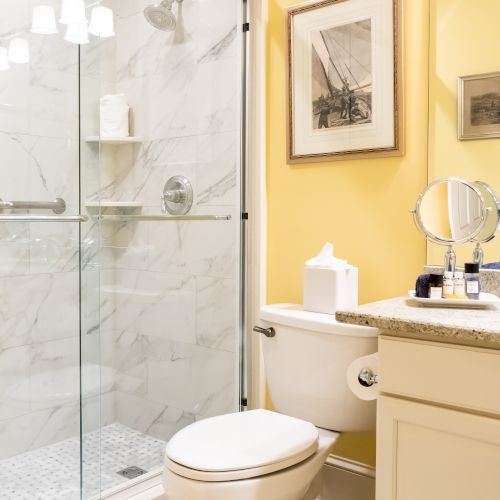 The image shows a bathroom with a toilet, a sink with a mirror, and a glass-enclosed shower next to it. The walls are painted yellow.