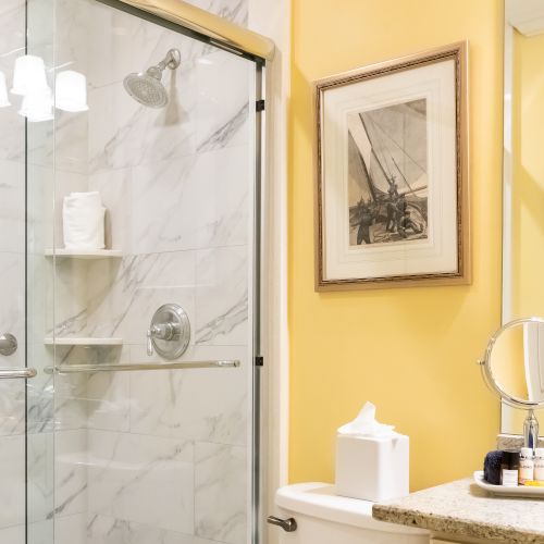 A bathroom with a glass shower, a vanity mirror, toiletries, tissue box, framed artwork, and a well-lit yellow wall.