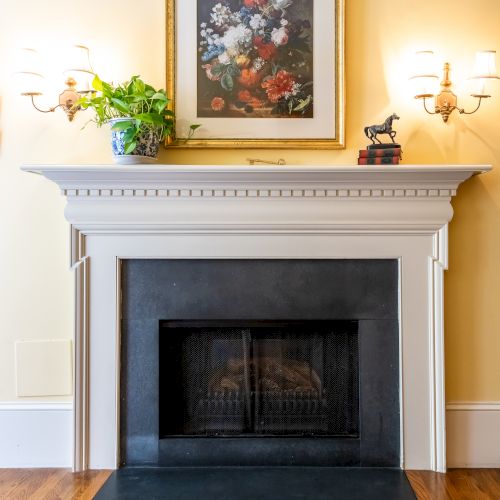 A classic fireplace with a black firebox, decorated with a framed floral picture, sconces, a plant, and a small sculpture on the mantel.