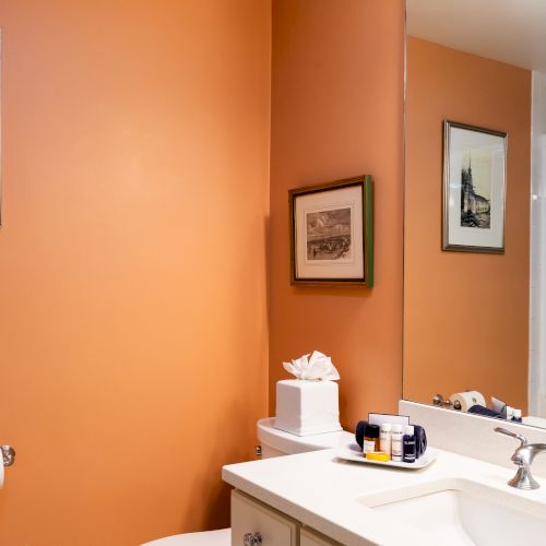 A bathroom with orange walls, mirror, sink, faucet, toilet, tissue box, framed pictures, toiletries, and a shower with sliding glass doors.