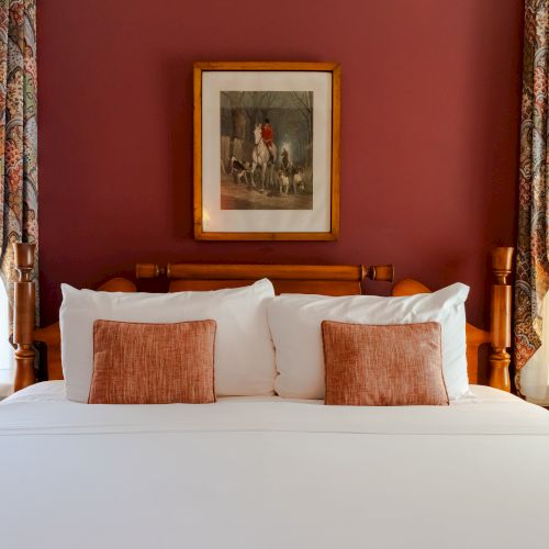 A neatly made bed with white linens and red accent pillows, flanked by two nightstands with lamps, and a framed picture on a maroon wall ends the sentence.